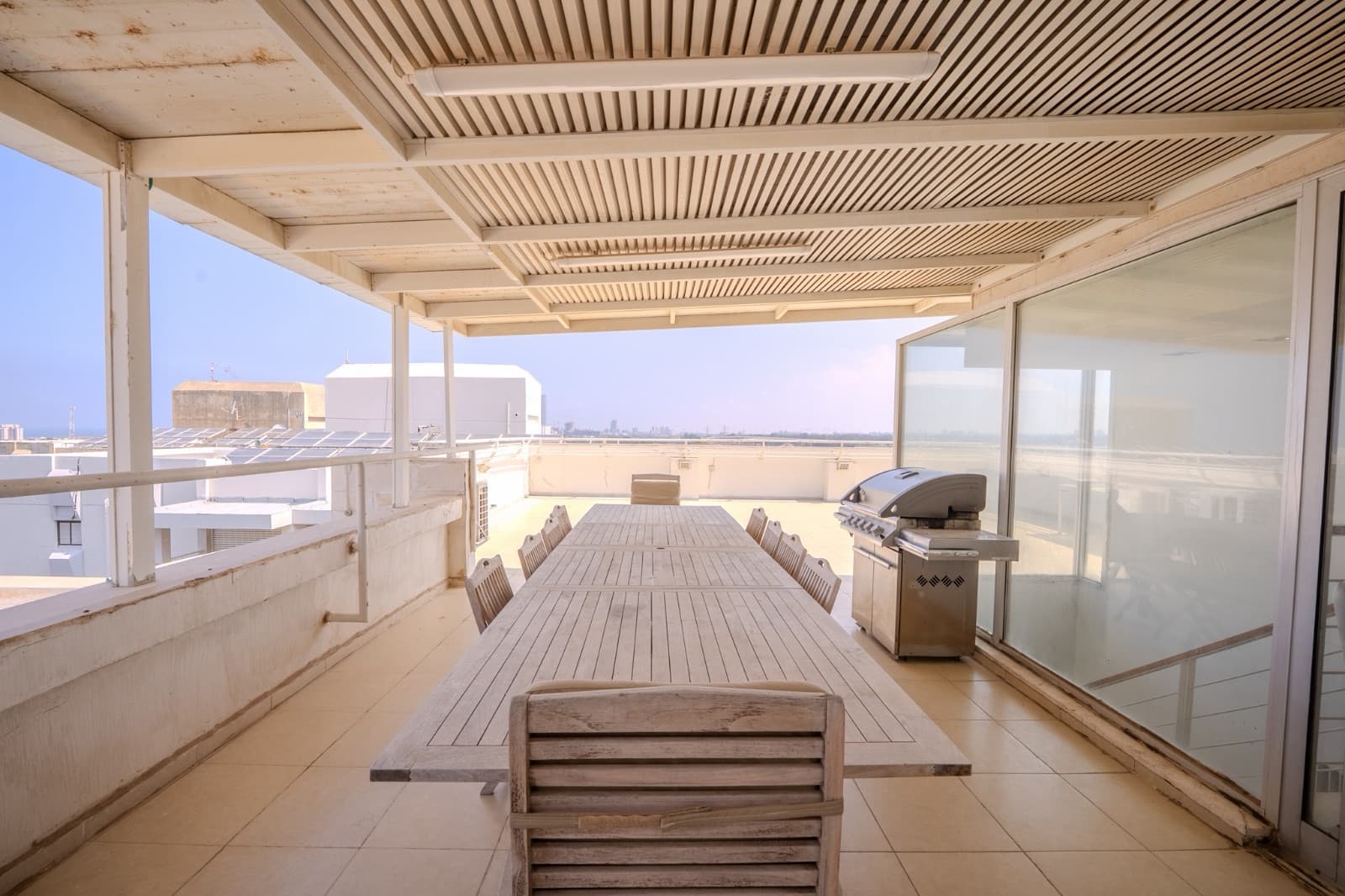 Covered rooftop dining terrace with a long teak table and barbecue under pergola slats